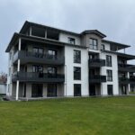 Die wichtigsten technischen Fensterparameter für Bürogebäude 10 Modernes Mehrfamilienhaus mit großen Balkonen, umgeben von gepflegtem Rasen und grauem Himmel im Hintergrund.