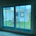 Warum sich neue Fenster und Garagentore in Berlin jetzt lohnen 4 Großes Fenster mit blauer Schutzfolie, Blick auf Baustelle und Garten im Hintergrund, moderne Architektur, Renovierung.