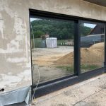PVC- vs. Aluminiumfenster: Welches Fenster für Ihr Gewerbeobjekt? 4 Große Glasschiebetür in einem Rohbau, Außenbereich mit Baustelle und Sandhaufen, Vorzeichen zukünftiger Fertigstellung.